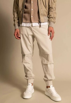 Homme portant un pantalon cargo beige avec des bas élastiques, des baskets blanches, et une superposition de veste, chemise et cardigan marron clair.
