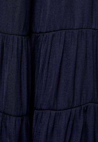 Tmavomodrý textil s jemnou, mierne pokrčenou štruktúrou. Obsahuje horizontálne vrstvené nariasenie vytvárajúce vrstvený vzhľad.