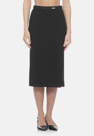 Donna che indossa una gonna a tubino nera al ginocchio con una cintura sottile e tacchi neri a punta, in piedi contro uno sfondo bianco.