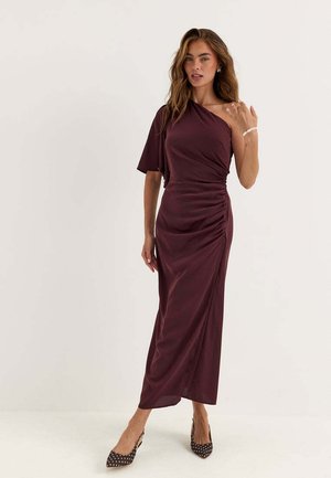 Donna che indossa un vestito arricciato monospalla color viola scuro con una manica e tacchi neri a punta con pois bianchi.