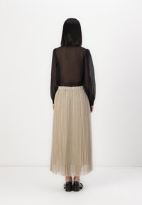 Svart gjennomsiktig bluse med lange ermer, kombinert med en beige plissert maxiskjørt med rynket midje og subtil glans. Svart fottøy fullfører antrekket.