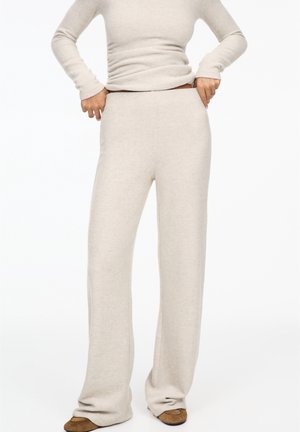 Persoon draagt beige gebreide broek en bijpassende top met lange mouwen, stelt de taille bij, staat met bruine schoenen op een witte achtergrond.