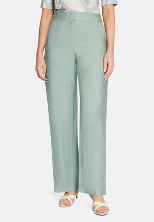 Kobieta ubrana w jasne zielone, szerokie spodnie z wysokim stanem, w połączeniu z białymi sandałami na obcasie z odkrytymi palcami, stojąca na jednolitym tle.