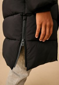 Čierna nafukovacia bunda s bočným zipsom, prešívaným dizajnom a manžetami s rebrovaním, kombinovaná so svetlo šedými nohavicami.