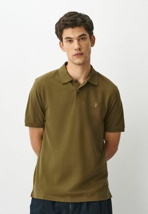 Ung mand med mørkt, krøllet hår iført en olivengrøn polo shirt med en lille hjorte-logo, står med hænderne bag ryggen.
