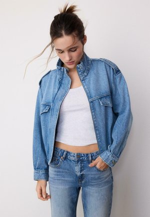 Ung kvinde med håret sat op iført blå denimjakke og jeans over en hvid cropped tanktop, kigger ned med den ene hånd i lommen.