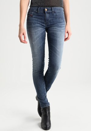 Niebieskie jeansy o dopasowanym kroju z subtelnym efektem przetarcia, noszone z czarnymi botkami, model stoi z nogami skrzyżowanymi na białym tle.