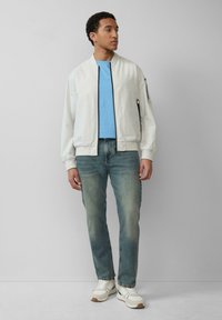 Witte bomberjack met zwarte rits, lichtblauwe t-shirt, vervaagde blauwe jeans en witte sneakers met beige accenten. Model staat zijwaarts.