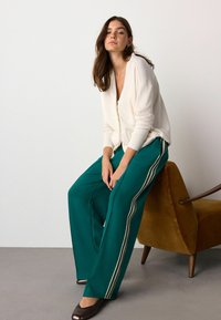 Weißer Cardigan, petrolfarbene weite Hose mit seitlichen Streifen und braunen High Heels. Modell sitzt auf einem braunen Akzentstuhl vor einem hellen Hintergrund.