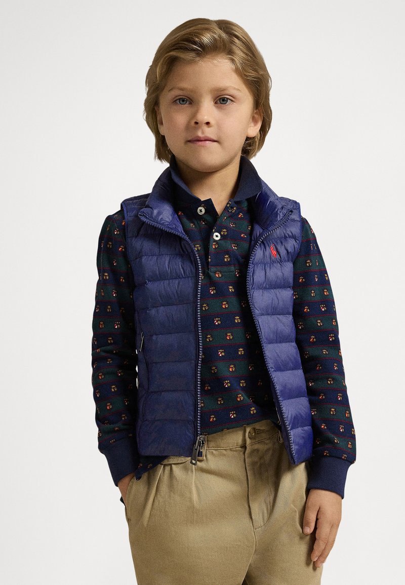 Marineblå vattert vest over en mørkegrønn skjorte med et lite blomsterprint, kombinert med beige bukser. Vesten har glidelås og sidelommer.