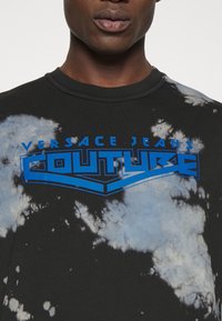 Schwarzes Batik-T-Shirt mit blauem "Versace Jeans Couture"-Logo. Weicher Stoff, normale Passform, runder Kragen und einzigartige wolkenartige Muster.