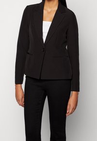 Czarna dopasowana marynarka z pojedynczym zapięciem na guzik, klapami, dwiema przednimi kieszeniami i długimi rękawami, w połączeniu z białą bluzką i czarnymi spodniami.