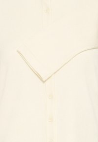 Cardigan en maille côtelée couleur crème avec fermeture à boutons, manches longues et texture douce. Détail de coutures visibles sur les bords.