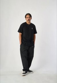 Camiseta V de algodón negra con pequeño logo en el lado izquierdo del pecho, combinada con pantalones negros holgados. Modelo vistiendo zapatillas negras, de pie sobre un fondo neutro.