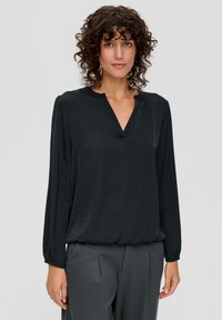 Schwarze Bluse mit V-Ausschnitt und langen Ärmeln, aus leichtem strukturiertem Stoff. Kombiniert mit grauen Hosen, glatter Oberfläche, lockere Passform.
