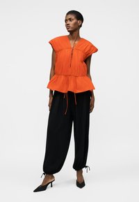 Femme portant un haut orange sans manches avec cordons et un pantalon noir ample avec des liens aux chevilles, debout en talons noirs pointus sur un fond blanc.