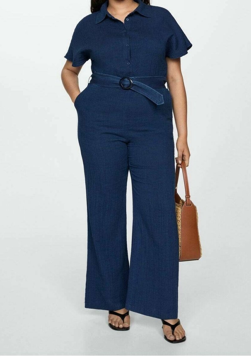 Femme portant une combinaison bleu marine ceinturée, à manches courtes et jambes larges, tenant un sac à main marron et portant des sandales noires.