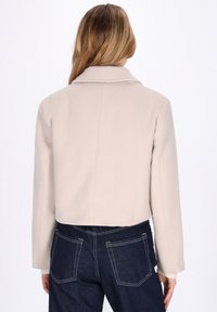 Beige cropped jakke med krave, strukturerede skuldre og lange ærmer. Bagsiden har en midterseam og ribbede manchetter. Sammen med mørke denimbukser.
