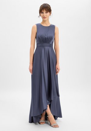 Femme portant une robe en satin bleu marine sans manches avec un corsage froncé et un ourlet asymétrique, associée à des sandales à talons hauts argentées, debout.