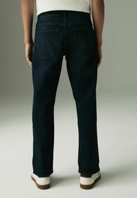 Tamsiai mėlynos džinsinės kelnės su tiesiu siluetu, su dviem galinėmis kišenėmis ir švariu apdailu. Nešiojamos su baltomis bateliais, esančiais neutraliame fone.