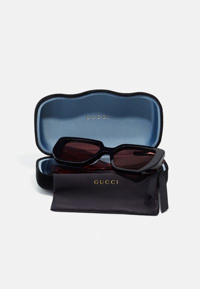Zwarte oversized zonnebril met donkere lenzen in een zwart en blauwe hoes, met een gouden "GUCCI" logo aan de binnenkant en een schoonmaakdoekje.