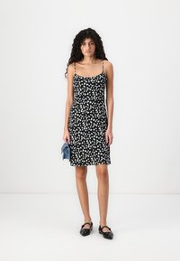 Rochie neagră cu un model abstract alb, cu bretele subțiri, lungime până la genunchi, design ajustat. Modelul ține o geantă albastră și poartă pantofi negri.
