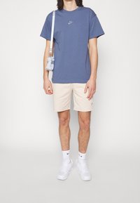 Camiseta de algodón azul de Nike con un pequeño logo, pantalones cortos beige, zapatillas blancas y una bolsa cruzada azul claro. Diseño atlético sencillo.