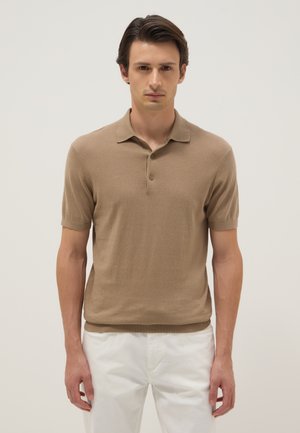 Mann mit dunklen Haaren, der ein beiges kurzärmeliges Polohemd und weiße Hose trägt, steht vor einem schlichten hellen Hintergrund.