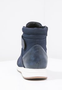 Mörkblå ankelstövlar med en texturerad bakdel, mjukt mockamaterial, vadderad krage och en kontrasterande vit sula. Detalj med kardborreband synlig.