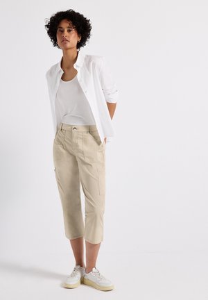 Pantalons cargo beiges avec poches, portés avec un débardeur blanc et une chemise blanche à boutons. Des baskets blanches complètent la tenue.