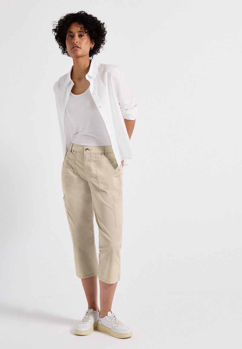 Pantaloni cargo beige con tasche, indossati con una canotta bianca e una camicia bianca con bottoni. Sneakers bianche completano l'outfit.