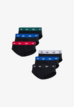 Mężczyzna bez koszulki, ubrany w czarne majtki z czarnym paskiem w talii ozdobionym białymi logo Adidasa, stojący z jedną ręką dotykającą szyi.