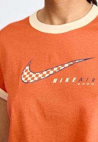 Oranje T-shirt met korte mouwen en crèmekleurige bies. Heeft een groot Nike Swoosh-logo in schaakpatroon en blauwe tekst "NIKE AIR ZOOM".