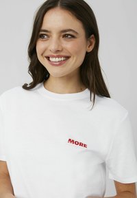 T-shirt en coton blanc à manches courtes. Présente un petit texte graphique rouge "MORE" et "pleasure" imprimé sur la poitrine. Encolure ronde.