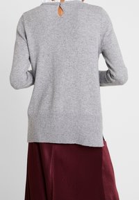 Grauer Strickpullover mit langen Ärmeln, geripptem Saum und einem Schlüssellochdetail am Rücken, über einem bordeauxfarbenen Satinenrock getragen.