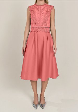 Robe midi corail sans manches avec corsage en dentelle, bords festonnés, jupe plissée, portée avec des escarpins argentés à bout pointu sur fond neutre.