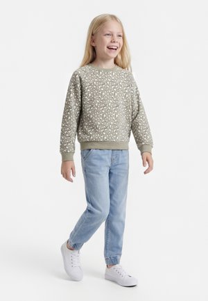 Niño sonriente con cabello largo y rubio camina, lleva un suéter gris claro con estampado de corazones, vaqueros azul claro y zapatillas blancas sobre fondo blanco.