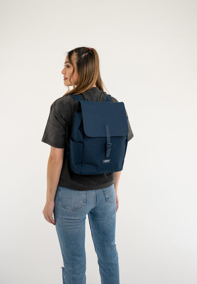 Mochila de tela azul marino con tapa con solapa, correas ajustables, cierre de hebilla y detalle del logo en la parte frontal. Material texturizado.