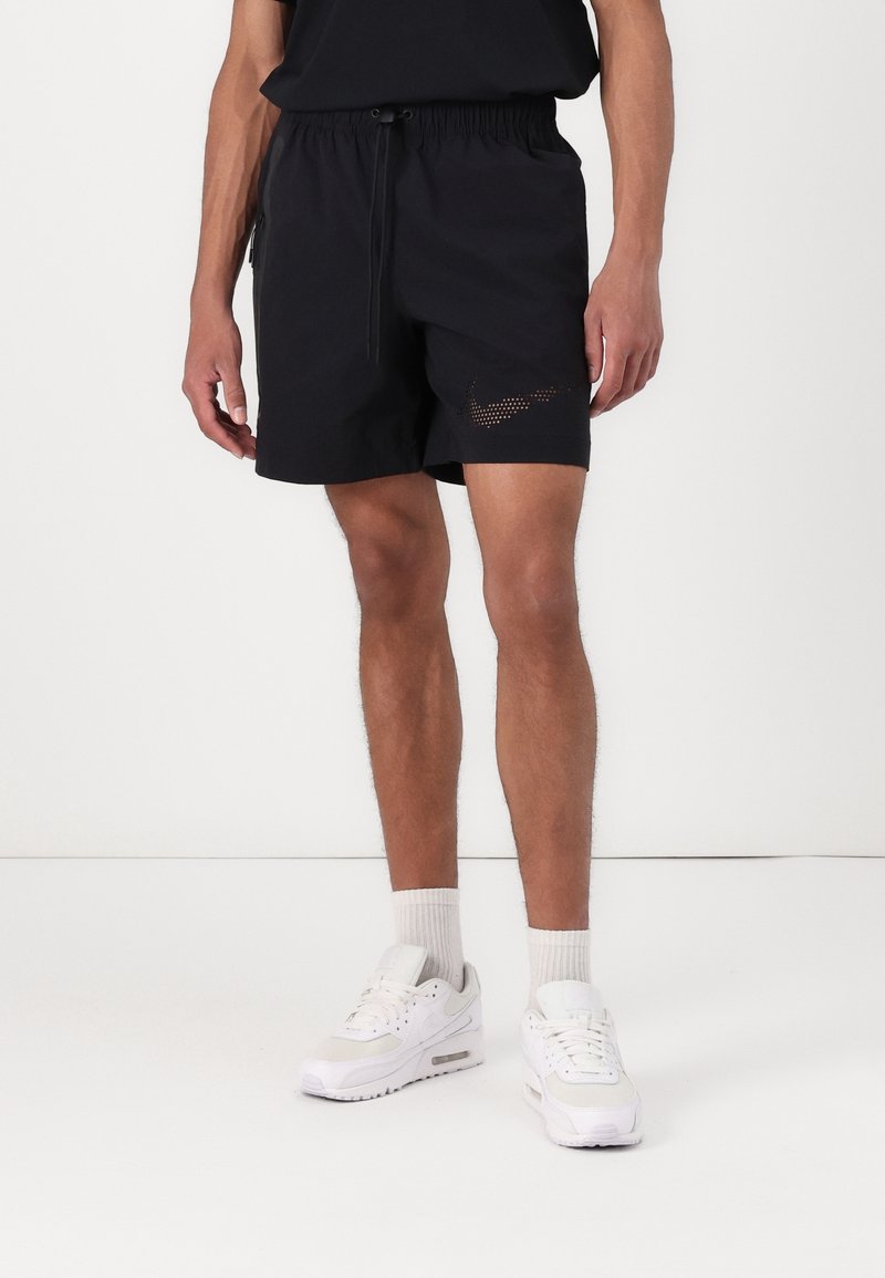 Homme portant un short de sport noir avec un logo swoosh en pointillé, des chaussettes blanches montantes et des baskets blanches sur un fond blanc uni.