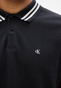 Polo noir avec doubles rayures blanches au col et logo "cK" blanc brodé sur la poitrine gauche.