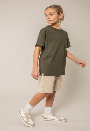 Enfant aux cheveux blonds portant un t-shirt vert olive, un short beige, des chaussettes blanches et des baskets blanches, marchant devant un fond neutre.