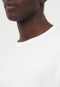 Κοντινό πλάνο του κάτω μέρους του προσώπου και του λαιμού ενός άνδρα που φορεί απλό λευκό μπλουζάκι με στρογγυλή λαιμόκοψη και με διακριτικό κεντημένο λογότυπο στο στήθος.