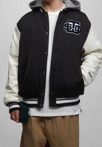 Czarna i kremowa kurtka bombery z szarym kapturem, paskowanymi mankietami, z przodu na zatrzaski oraz z haftowanym logo na piersi.