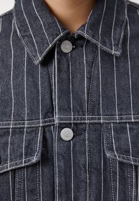 Jeansjack met een marineblauwe basis en witte verticale strepen, voorzien van een puntige kraag, dubbele borstzakken en metalen drukknoopsluitingen.