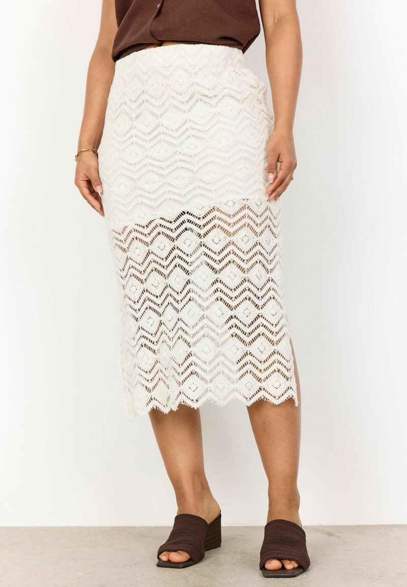 Jupe midi en dentelle blanche avec un motif en zigzag, présentant une partie inférieure transparente et une taille texturée. Associée à des sandales à plateforme brown.