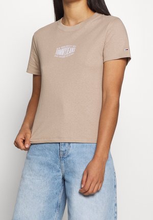 Kvinde iført en beige Tommy Jeans t-shirt med rund hals og lyseblå højtsiddende jeans, stående mod en hvid baggrund.