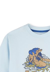 Lichtblauwe katoenen T-shirt met een grafiek van bruine laarzen met blauwe veters op een witte spetter, omlijnd in het geel. Ronde halslijn, korte mouwen.