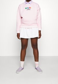 Sudadera rosa con logo bordado, falda blanca y zapatillas de cuadros morados. Superpuesta con una camisa ligera de color púrpura con cuello.