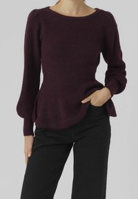 Frau in einem dunkelvioletten Strickpullover mit gepufften langen Ärmeln und schwarzer Hose, mit einer Hand in der Tasche, vor einem einfarbigen Hintergrund.