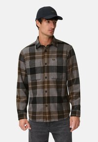 Flannel shirt in gray, black, and brown plaid with two front pockets and button closure. Paired with a navy cap.